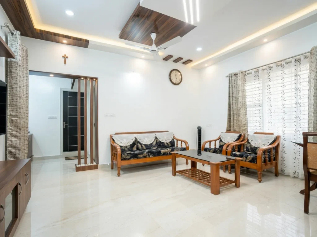 Spacious living room interior featuring a luxury false ceiling with wooden accents, vitrified flooring, and traditional wooden furniture.