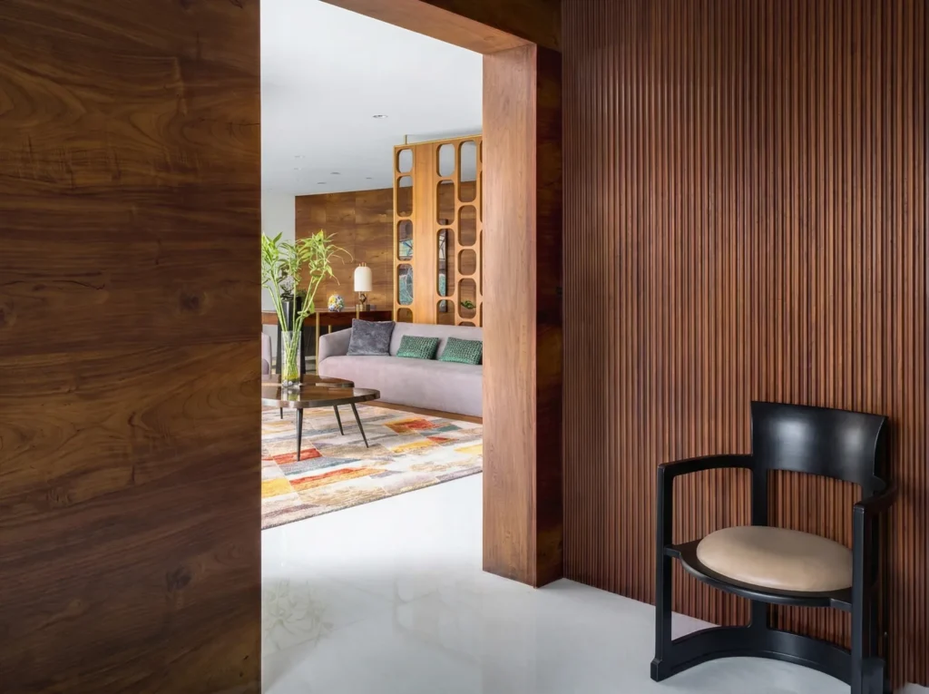 Arun Sky Garden residence – foyer entry with vertical teak fluted wall panelling, barrel accent chair, and view into furnished living room by High Tech Constructions, Bangalore