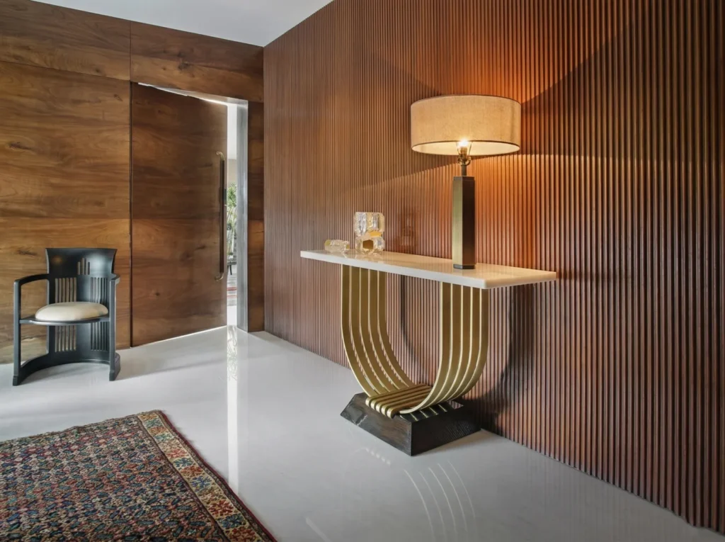 Arun Sky Garden residence – entryway with fluted teak wall panel, marble-top gold arc console table, statement table lamp, and full-height pivot door by High Tech Constructions, Bangalore
