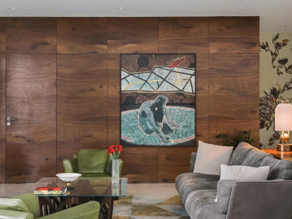 Arun Sky Garden residence – living room feature wall with book-matched walnut veneer panelling, large contemporary artwork, grey sofa, and green leather accent chairs by High Tech Constructions, Bangalore