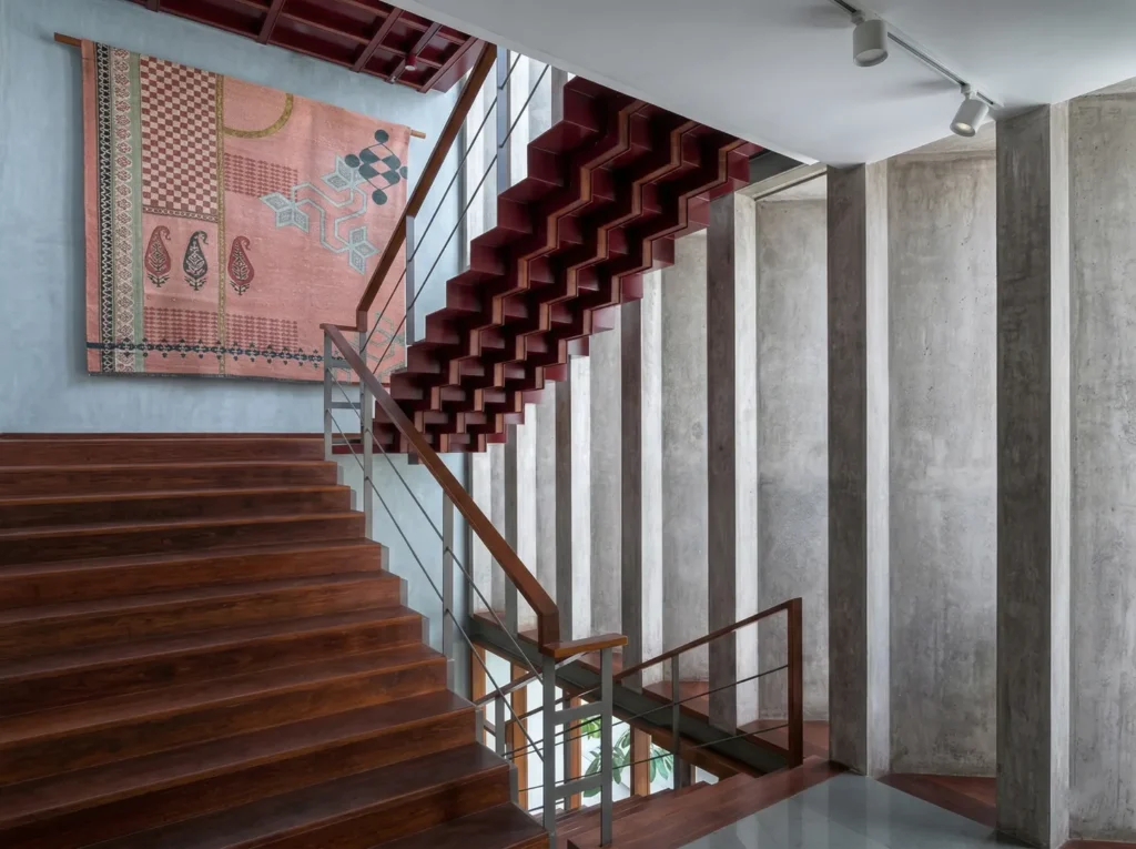 Double-height staircase hall with crimson floating stairs, exposed concrete, and traditional textile wall art in a Bangalore home construction