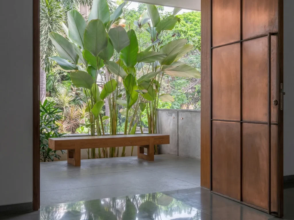 Residential home entrance in Bangalore with a copper pivot door opening to a lush tropical garden courtyard by High Tech Constructions