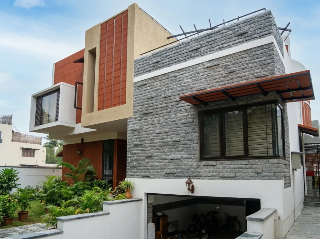 Kalpana turnkey bungalow – exterior side view featuring grey granite stone cladding, terracotta tile facade, wooden pergola canopy, and basement garage by High Tech Constructions, Bangalore