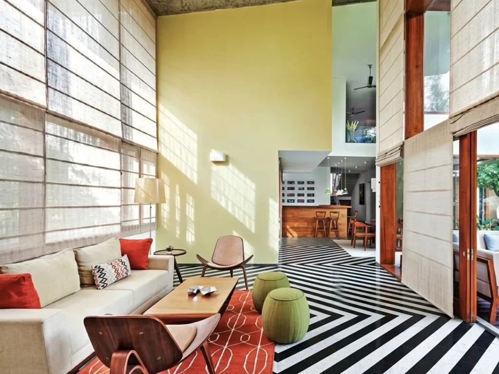 Vibrant double-height residential living room interior in Bangalore with striped stone floor, burlap shades, and open kitchen view by High Tech Constructions