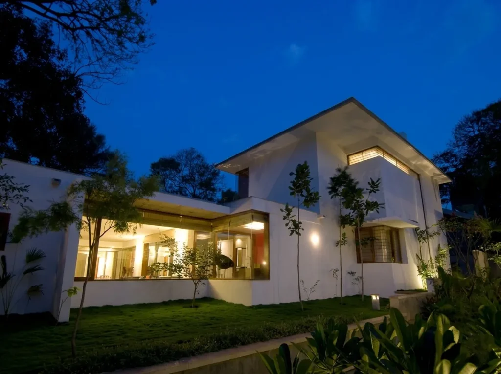 Modern residential home construction in Bangalore glowing at night with full-width glazing, white volumes, and landscaped garden by High Tech Constructions