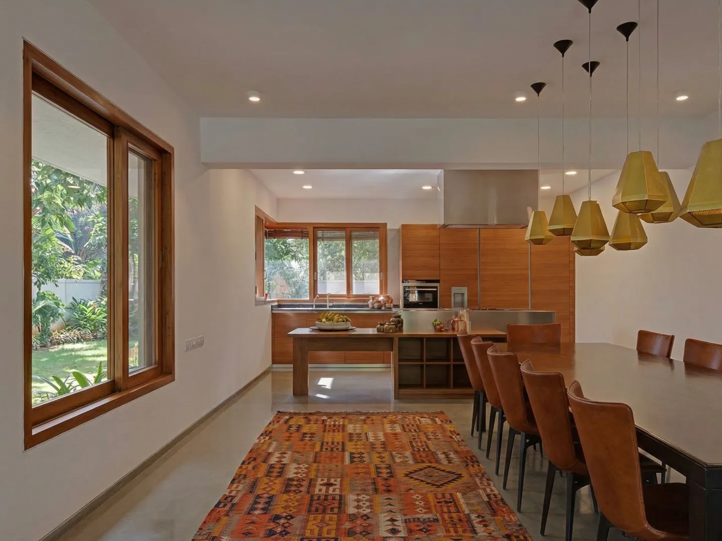 Open-plan kitchen and dining interior construction in Bangalore with timber cabinetry, bamboo pendant lights, and garden window view by High Tech Constructions