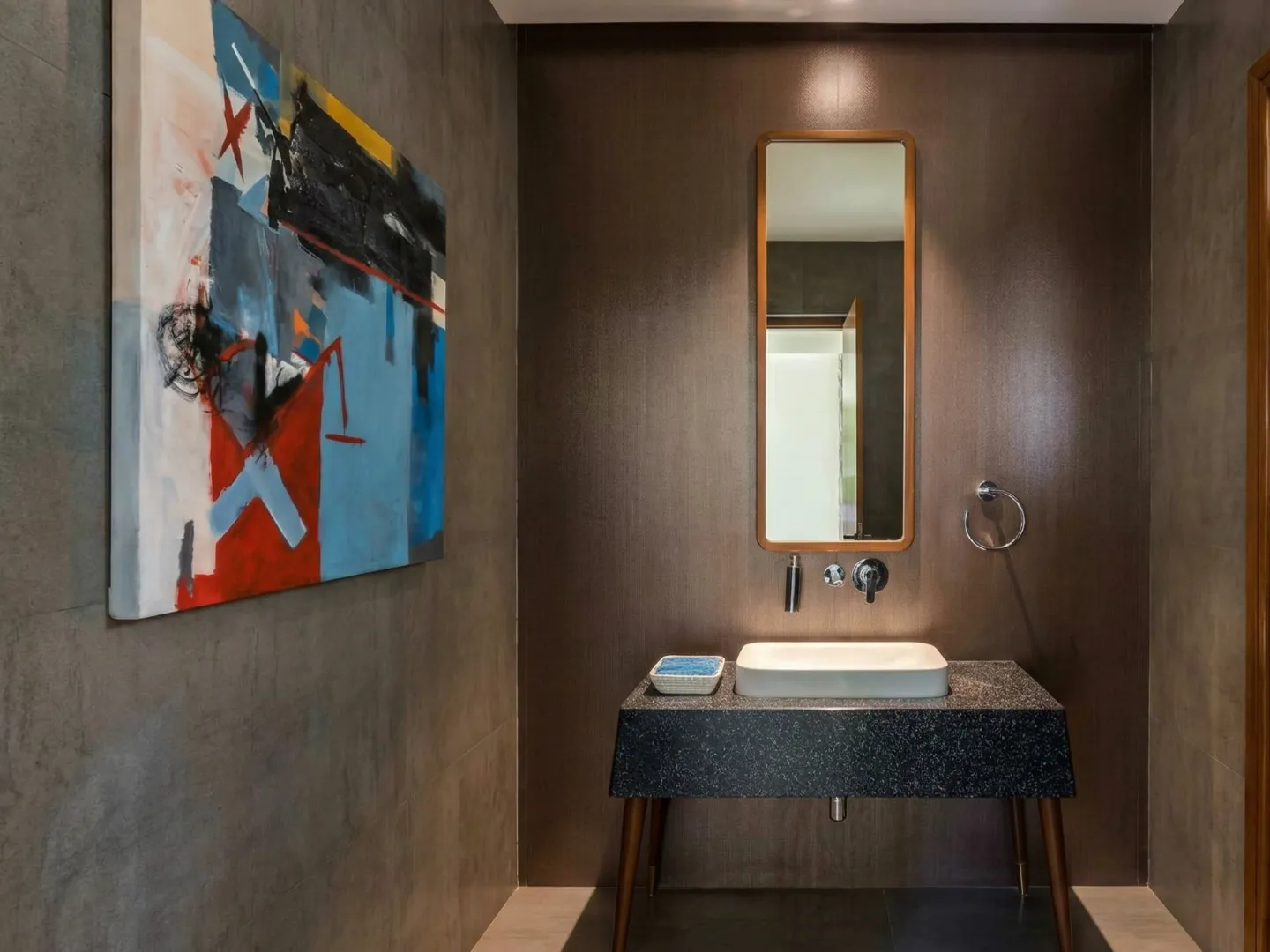Designer powder room interior construction in Bangalore with terrazzo vanity, teak-framed mirror, dark timber walls, and abstract artwork by High Tech Constructions