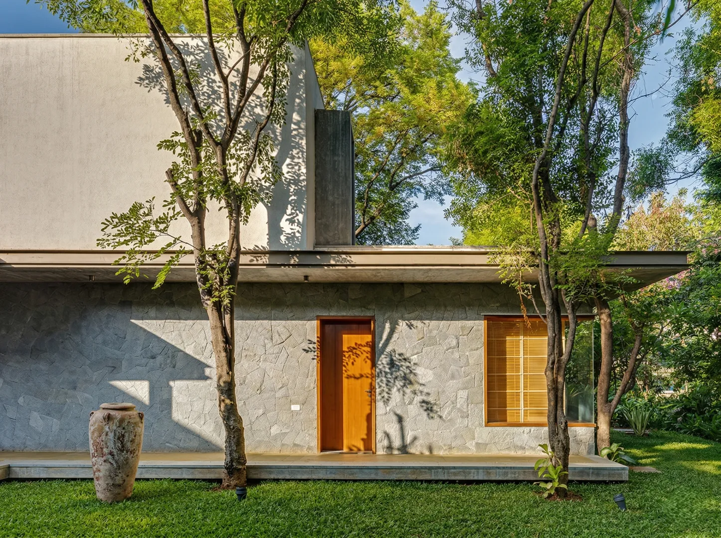 Residential home construction in Bangalore with rough-dressed granite stone cladding, teak entry door, flat cantilevered roof, and dappled tree light by High Tech Constructions