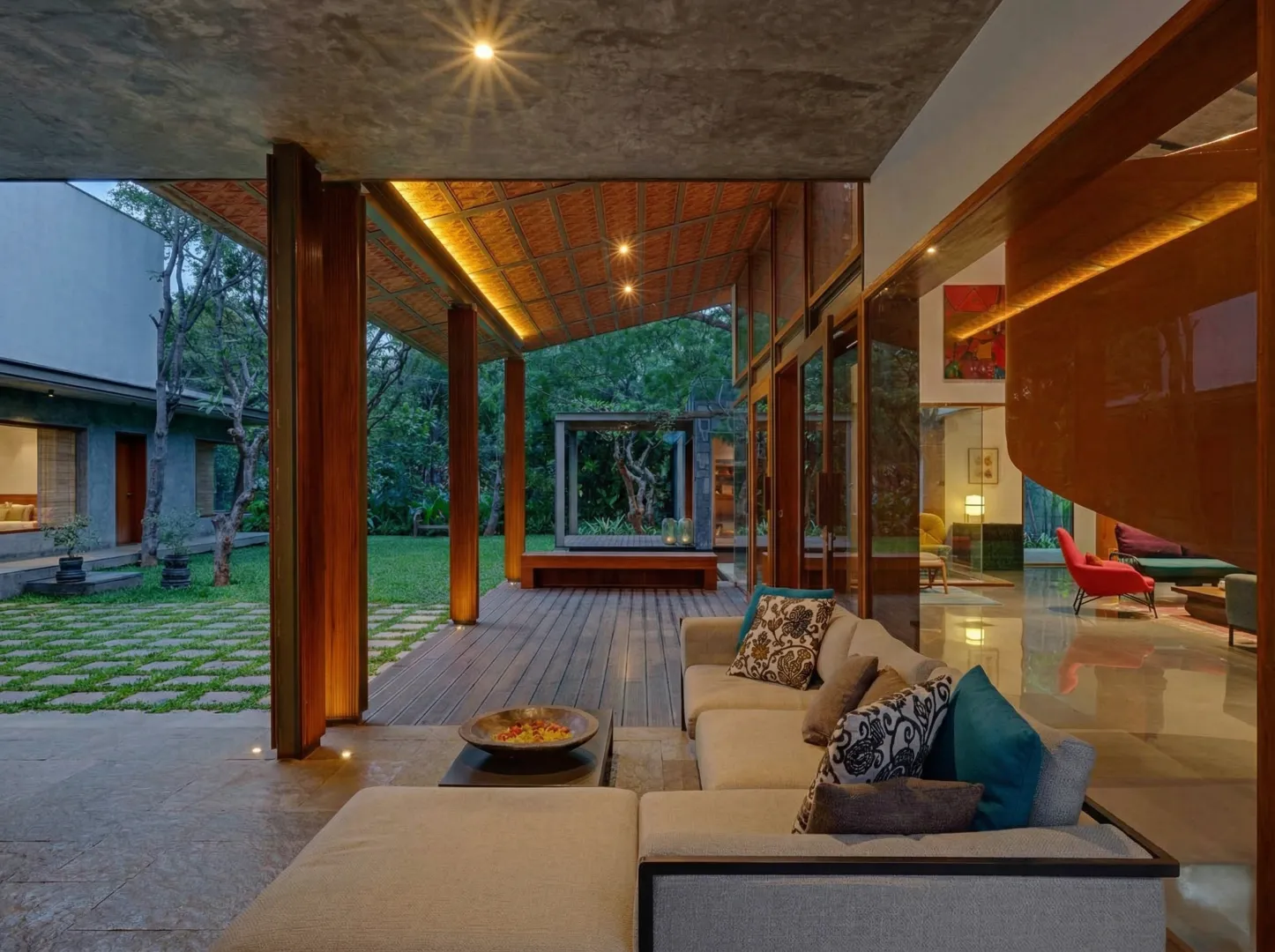 Residential covered outdoor verandah with terracotta tile ceiling, teak columns, sectional sofa, and courtyard garden view at dusk in a Bangalore home construction