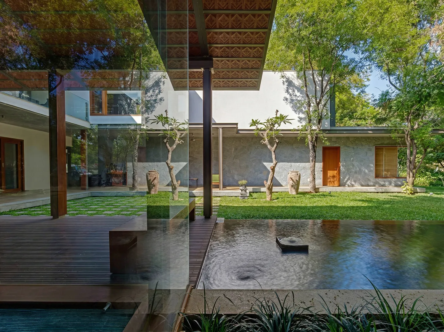 Residential home construction in Bangalore showing stone-clad wing, central lawn garden, and glass reflection of terracotta roof by High Tech Constructions