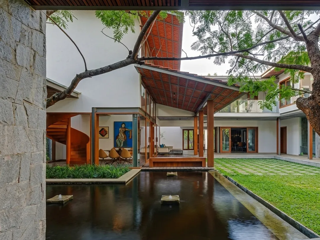 Wide residential courtyard view in Bangalore with reflecting pool, terracotta gabled roof, spiral staircase glimpse, and mature tree canopy by High Tech Constructions