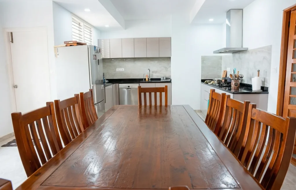 Open-plan dining room with solid wood table and modular kitchen in a residential construction project in Bangalore.