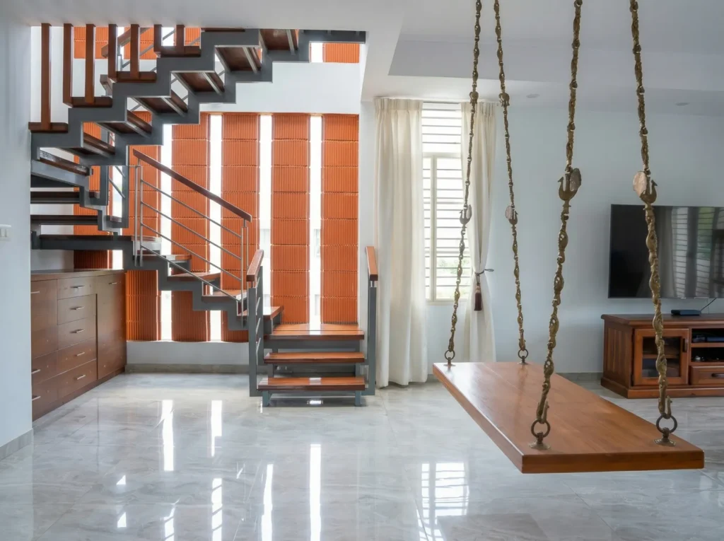 Residential living room with floating open-riser staircase, terracotta brick wall, and traditional wooden indoor swing — home construction Bangalore.