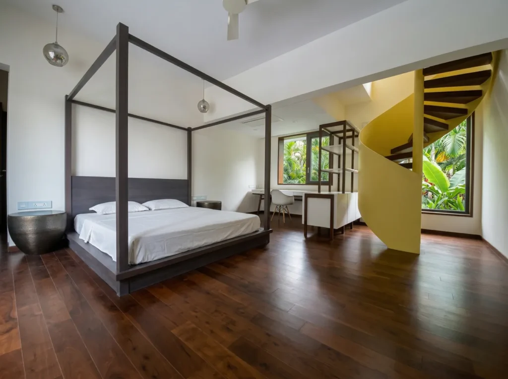 Premium master bedroom with four-poster teak bed, hardwood flooring, and yellow spiral staircase opening to tropical garden — home construction Bangalore.