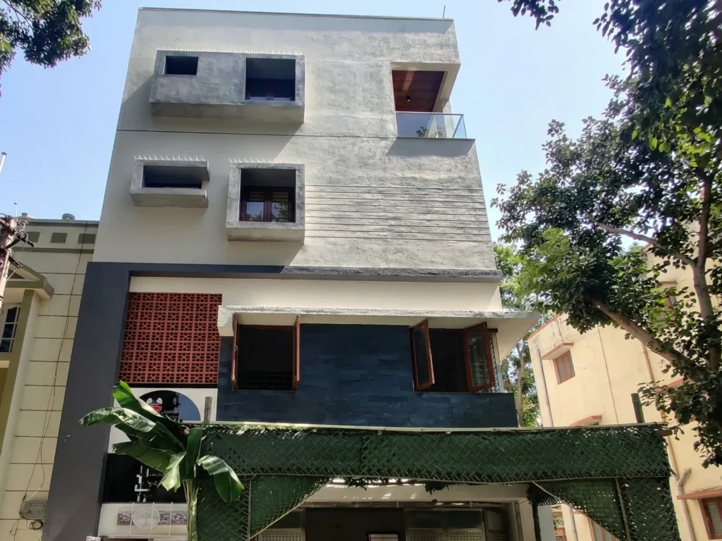 Contemporary multi-story residential elevation featuring a minimalist grey cement finish, decorative terracotta jaali work, and dark slate stone wall cladding by High Tech Constructions.