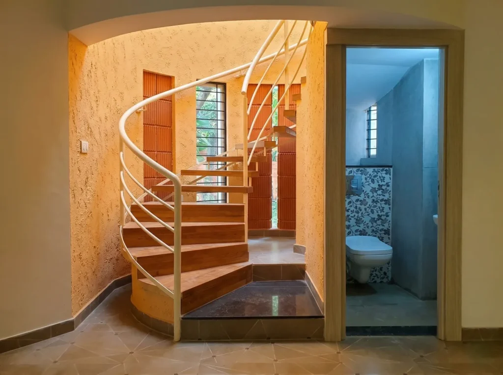 Interior wooden spiral staircase with white railings and natural lighting through architectural terracotta slits and textured orange walls.