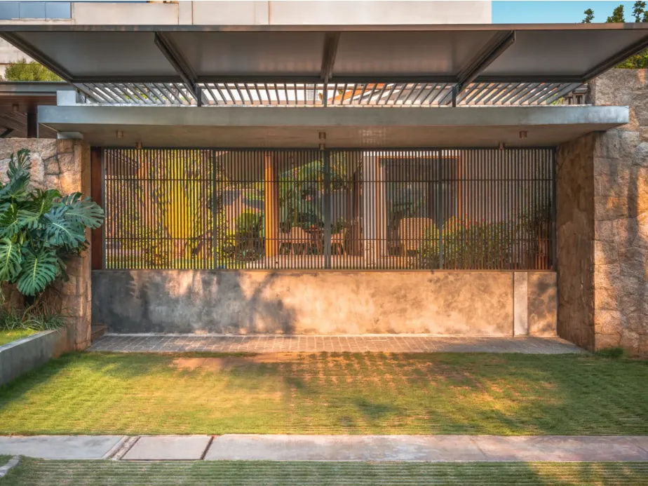 Residential front entry gate with rough granite stone cladding, teak vertical louvre screen, steel pergola canopy, and manicured lawn in a Bangalore home construction by High Tech Constructions