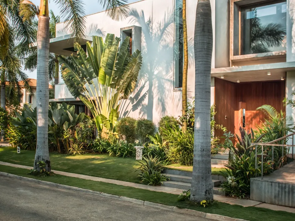 Modern residential home construction in Bangalore with white rendered facade, large-format glazing, teak entry door, and lush tropical street garden by High Tech Constructions