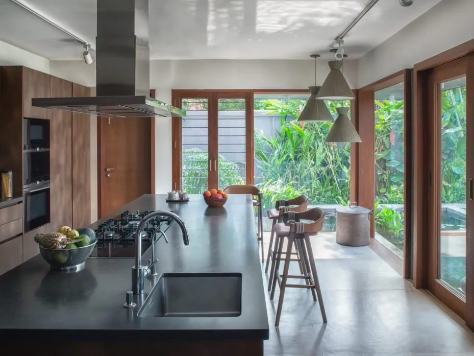 Modern residential kitchen interior construction in Bangalore with black granite island, walnut cabinetry, bar stools, and tropical garden view by High Tech Constructions