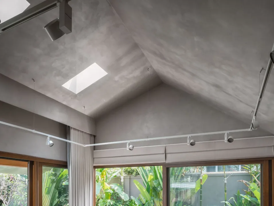 Residential interior with vaulted grey micro-cement ceiling, central skylight, track lighting, and full-height teak-framed garden windows in a Bangalore home construction