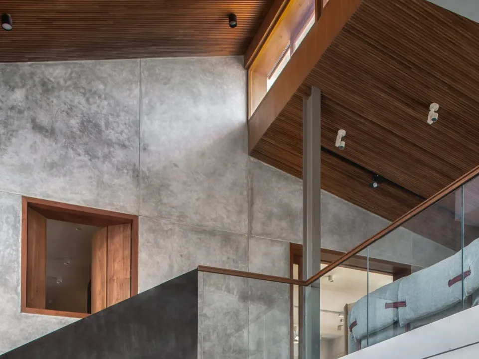 Double-height residential staircase void with micro-cement walls, warm teak timber ceiling, glass balustrade, and teak-framed internal window in a Bangalore home construction