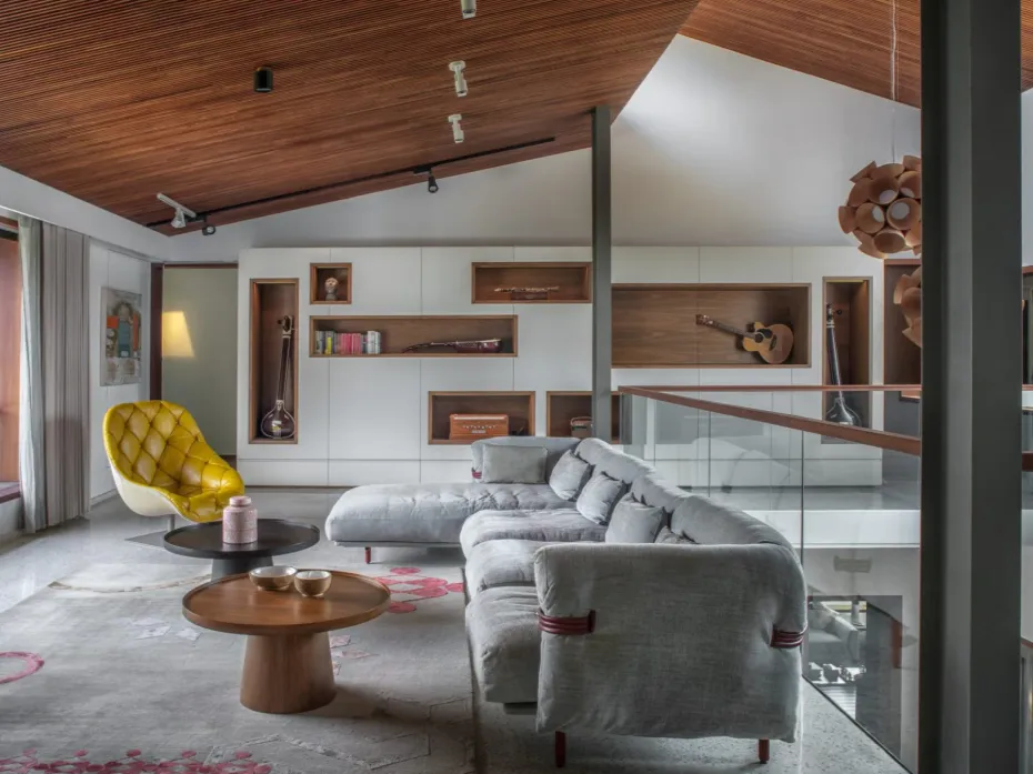 Residential upper-level family room interior in Bangalore with teak timber ceiling, musical instrument display wall, grey sectional sofa, and yellow accent chair by High Tech Constructions
