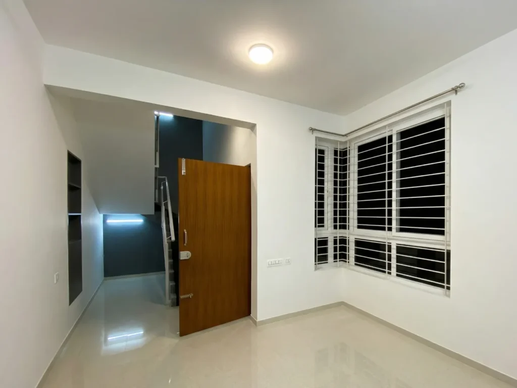Interior view of a modern apartment featuring a solid wood main door, large corner windows with safety grills, and high-gloss flooring.