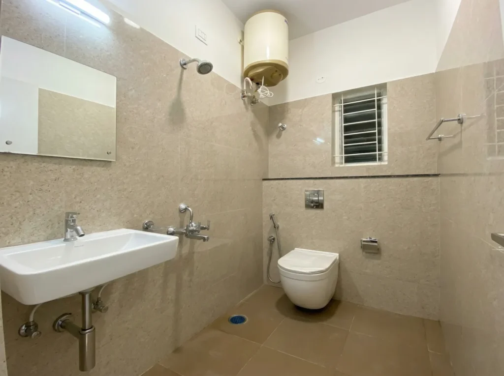 Spacious master bathroom featuring beige stone-finish tiles, a wall-mounted washbasin, and a professional plumbing installation with geyser