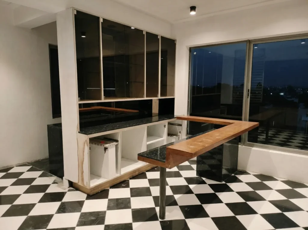 Residential bar unit under construction in Bangalore with black granite countertop, teak bar top, dark glass cabinetry, and checkered marble floor by High Tech Constructions