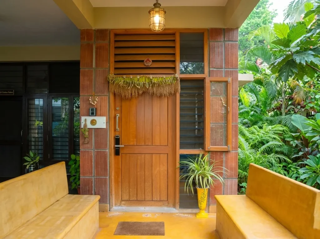 Anand Prakash residence – main entrance with teak wood door, clay brick surround, traditional cage pendant light, yellow oxide seating, and garden frontage by High Tech Constructions, Bangalore