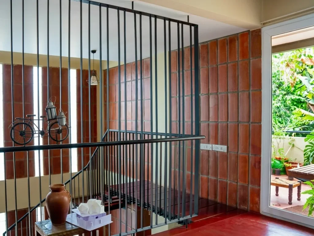 Anand Prakash residence – upper floor landing with red oxide flooring, MS vertical bar railing, clay tile wall cladding, and balcony garden view by High Tech Constructions, Bangalore