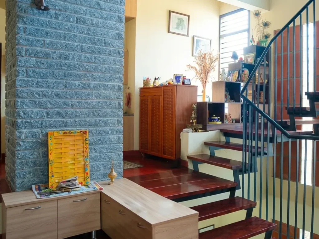 Anand Prakash residence – living area with granite stone-clad column, MS staircase railing, red oxide flooring, and traditional wooden cabinetry by High Tech Constructions, Bangalore