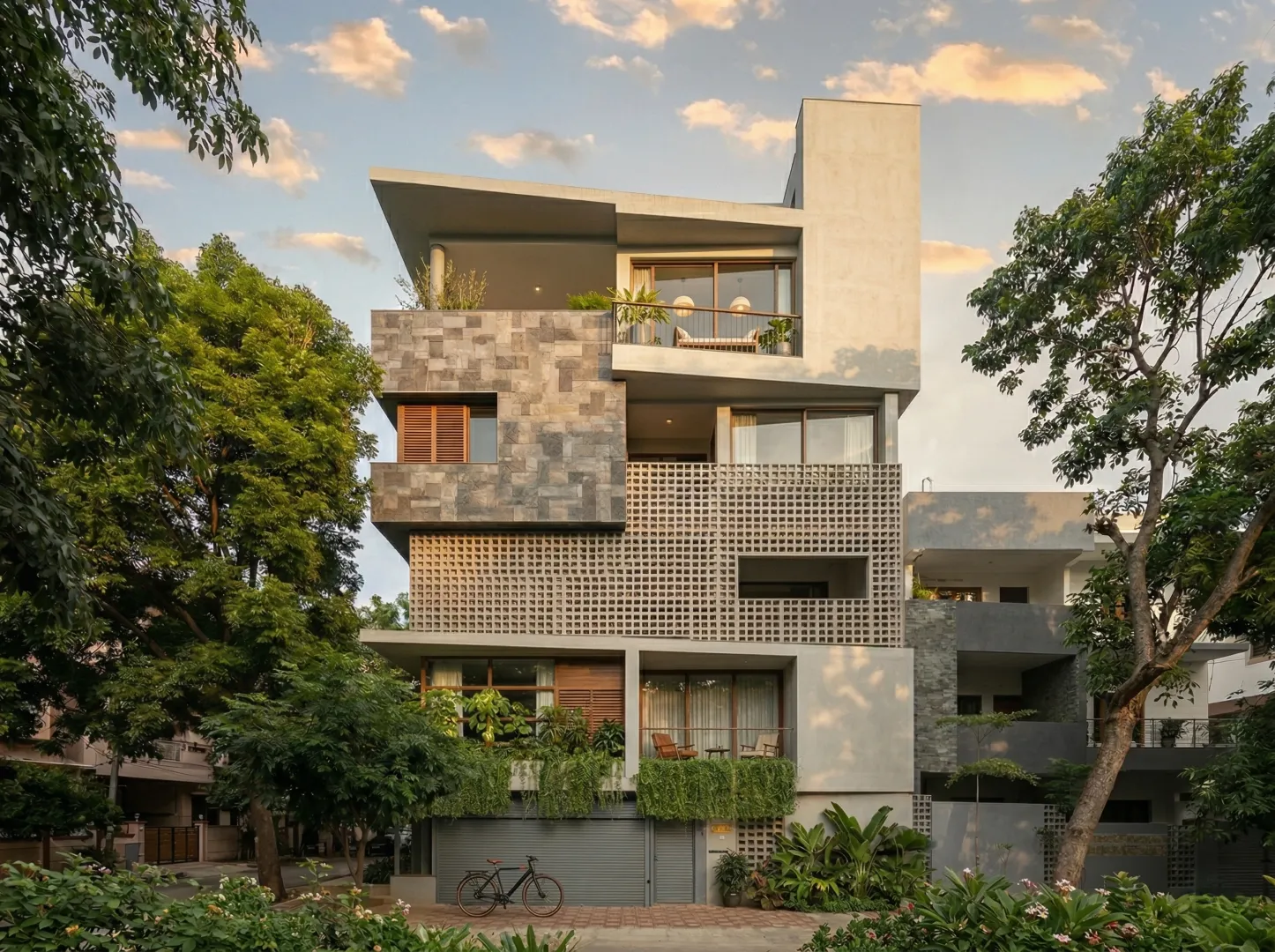 Arun Charanthimath residence – sunset view of front elevation featuring stone mosaic upper floor, concrete jali screen, green balconies, and rooftop garden by High Tech Constructions, Bangalore