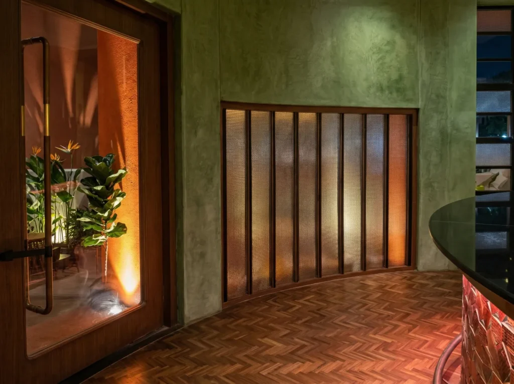 Restaurant interior construction lobby in Bangalore with herringbone timber floor, sage green walls, and warm backlit glass panels by High Tech Constructions