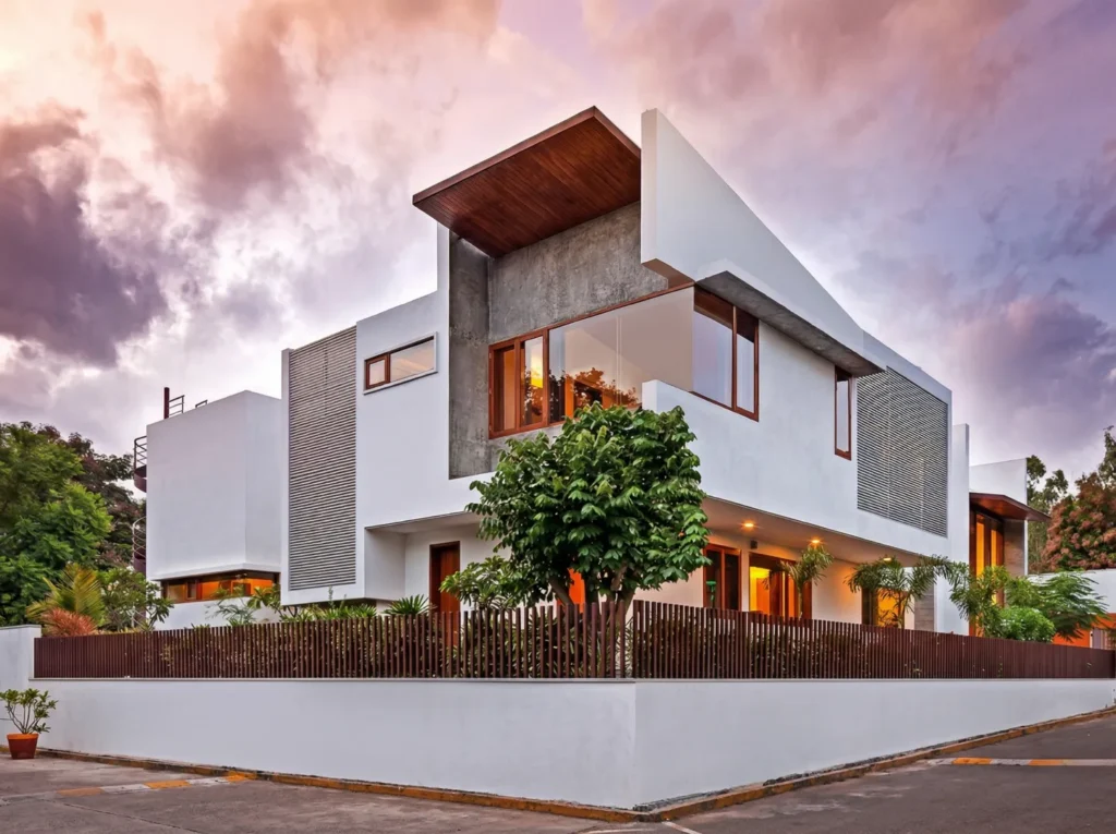 Modern residential construction in Bangalore with cantilevered timber roof, full-height glazing, garden deck, and outdoor lounge at twilight