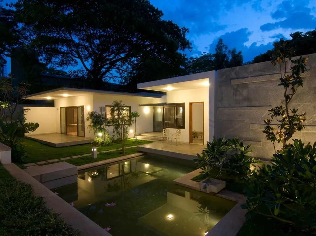 Residential home construction in Bangalore with illuminated garden pavilion, reflecting pool, granite boundary wall, and tropical planting at dusk