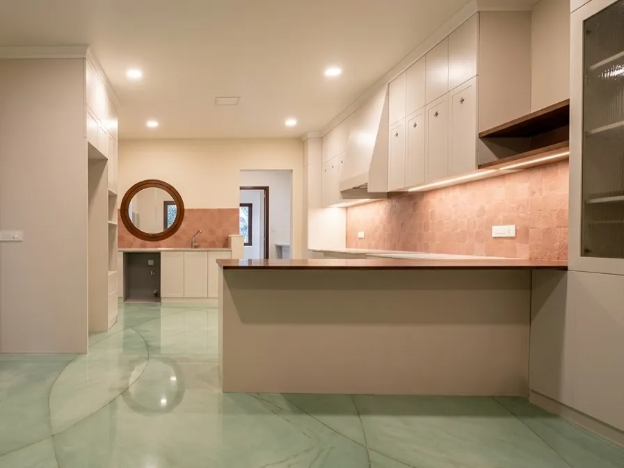 Modern kitchen with white modular cabinets, timber island countertop, pink stone backsplash, and green terrazzo flooring — home construction Bangalore.