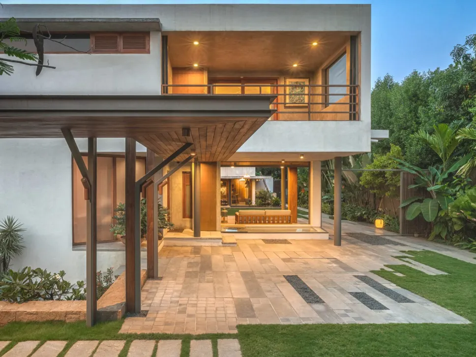 Residential covered driveway pavilion with teak ceiling, steel columns, stone-paved forecourt, and warmly lit interior at dusk in a Bangalore home construction by High Tech Constructions