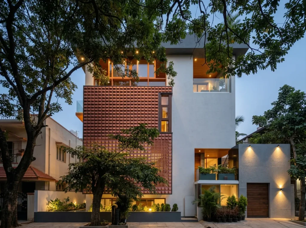 Modern multi-storey home construction in Bangalore with terracotta jaali screen facade, teak ceiling details, and landscape lighting at dusk by High Tech Constructions
